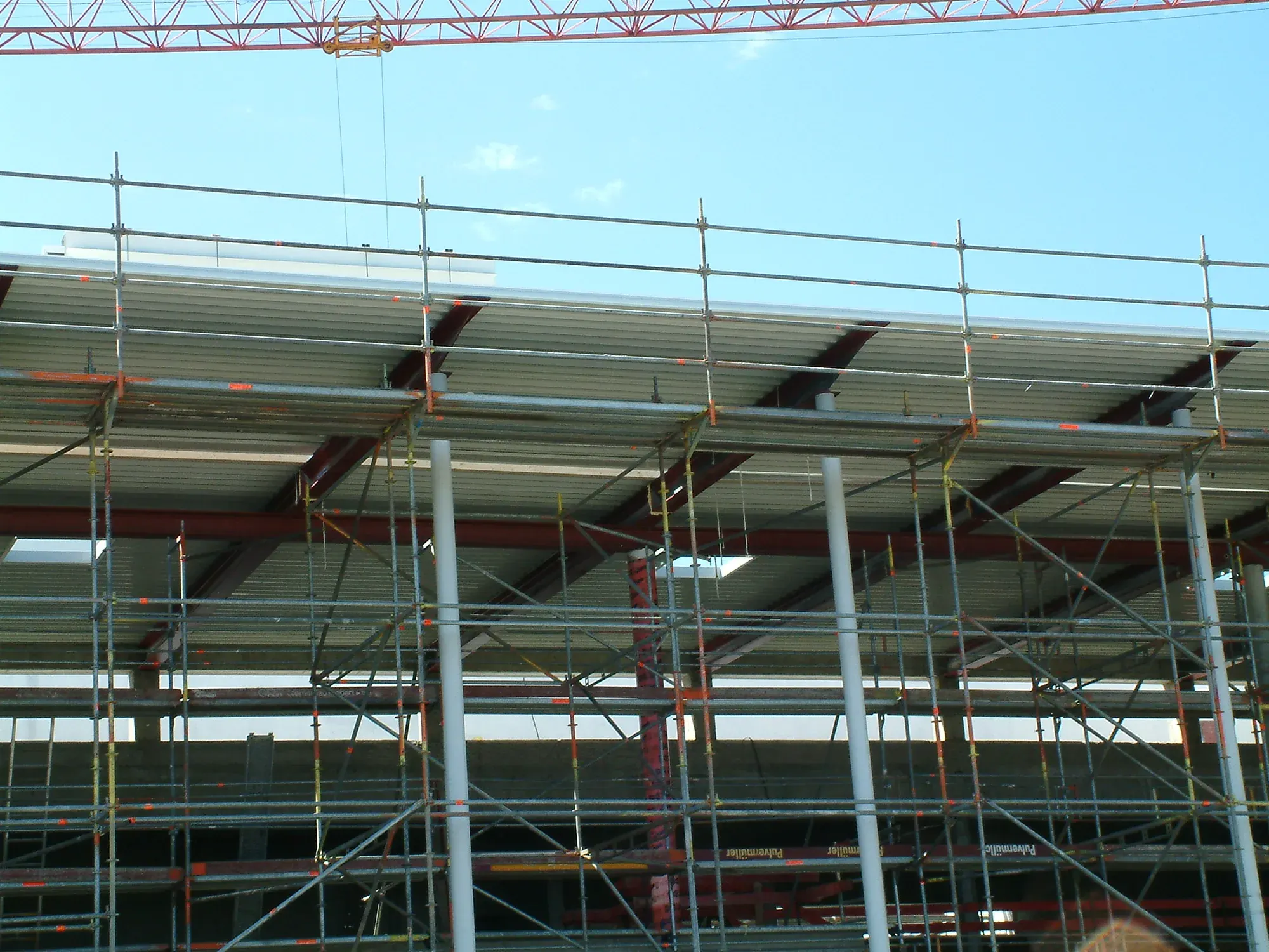 Rohbau eines langgestreckten Multiplexgebäudes mit eingerüsteter Fassade und weit auskragendem Vordach vor blauem Himmel